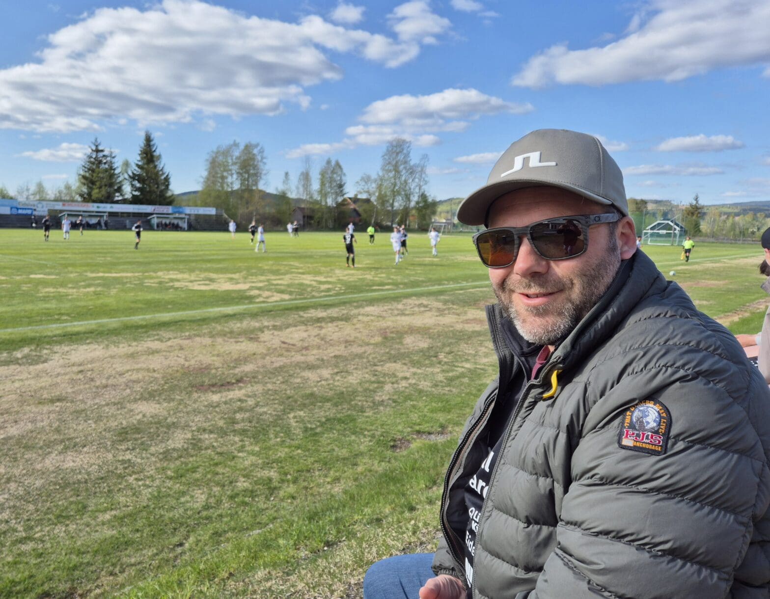 Hans Otto Veflen smiler på fotballbane under klar blå himmel, iført caps, solbriller og boblejakke, under Odal FKs første hjemmekamp.