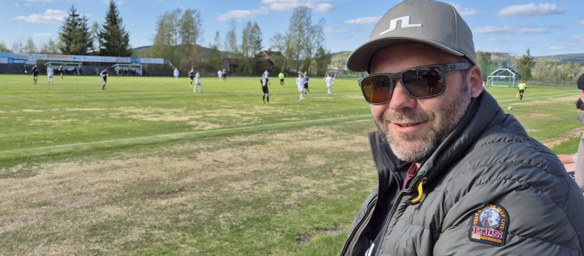 Hans Otto Veflen smiler på fotballbane under klar blå himmel, iført caps, solbriller og boblejakke, under Odal FKs første hjemmekamp.