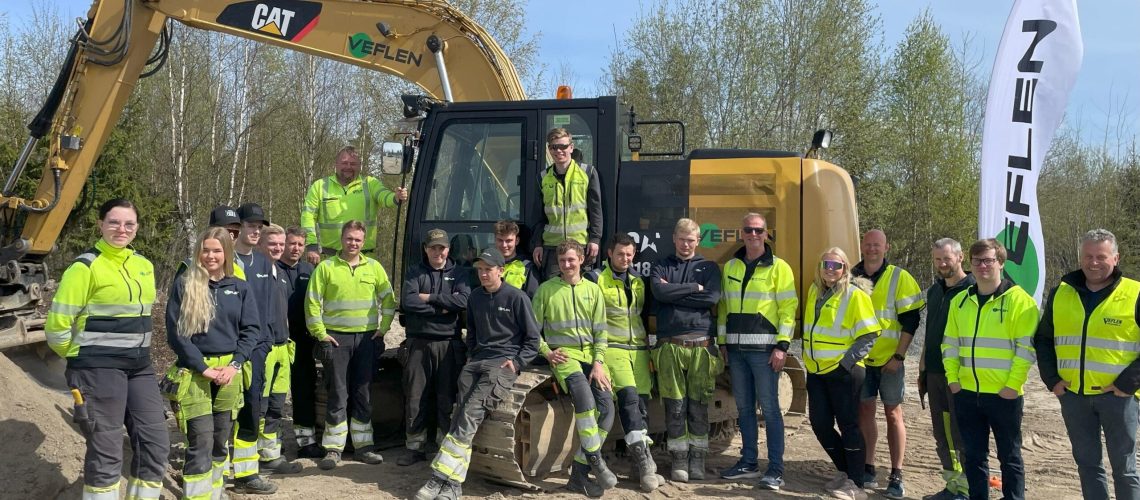 Gruppebilde av lærlinger, mentorer og ledere foran anleggsmaskiner, tatt under en vellykket lærlingedag i vårsola hos Veflen.